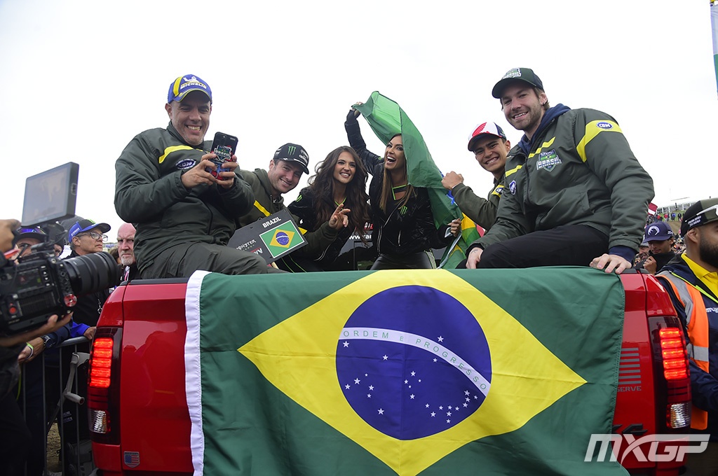 Apresentação do time Brasil no Motocross das Nações 2024 no circuito de Matterley Basin, na Inglaterra. Em cima da caminhonete vermelha temos da esquerda para a direita: Cali Neto chefe de equipe mxon Brasil 2024 e os pilotos brasileiros Fábio Santos da categoria Open, Bernardo Tibúrcio da categoria MX2 e Enzo Lopes da categoria MXGP. Duas garotas monsters girls em cima da caminhonete agitando a bandeira do Brasil para animar a torcida, uma monster girl é a brasileira Ana Carolina Benitez.