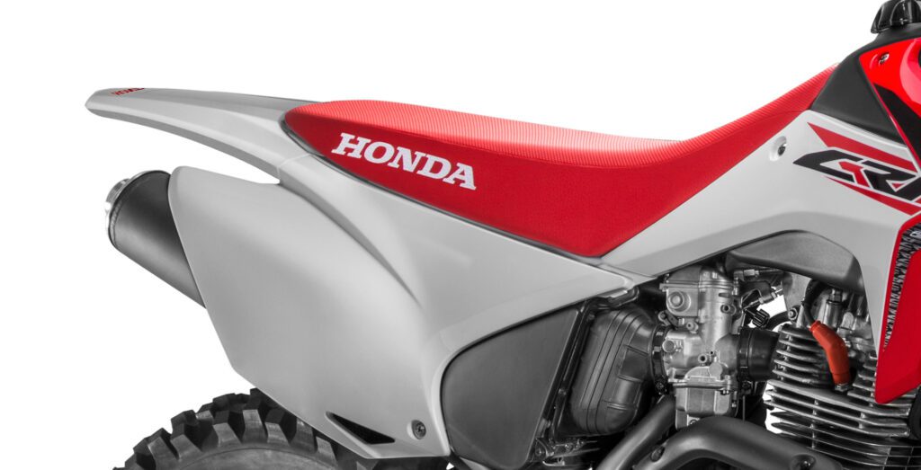fotografia da moto honda crf 230 do lado direito mostrando a capa de banco e assento, paralama traseiro, tanque de combustível. escapamento completo cano e ponteira, carburador original e uma parte do motor